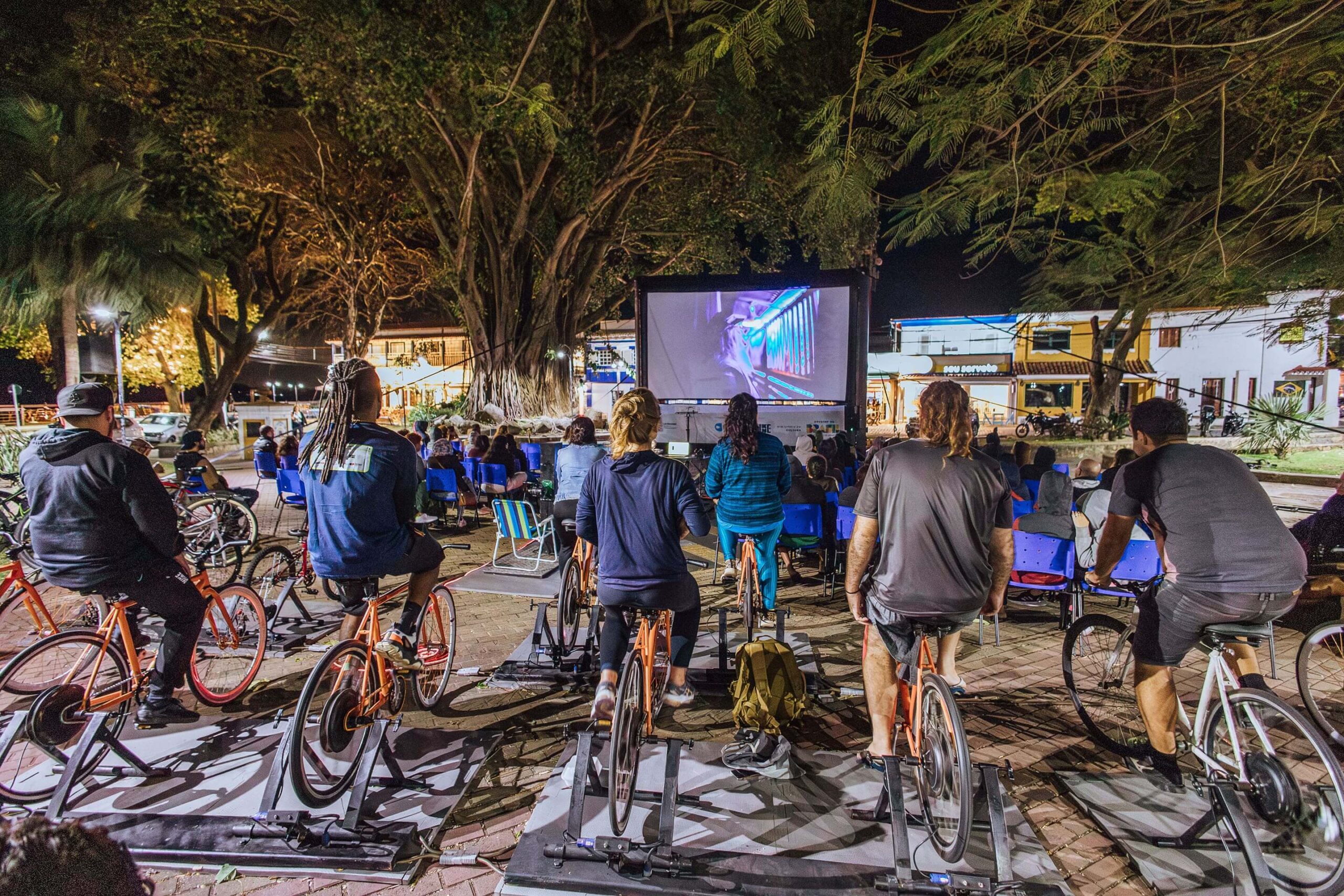 Fotografia em plano geral, retratando um evento de cinema ao ar livre em uma praça pública. Em primeiro plano, pessoas de costas pedalam bicicletas laranjas fixas enquanto assistem a um filme na tela grande à frente. Ao fundo, árvores grandes e casas iluminadas.
